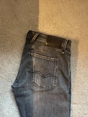 Replay Jeans W31 - Säljer ett par grå jeans från Replay med slitningar och distressed detaljer på både fram- och baksida. Jeansen har klassisk femficksmodell, raka ben och en snygg tvättad look. Perfekta för dig som gillar en avslappnad och trendig stil.