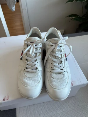 Vita Gucci sneakers med röd-blå rand - Snygga vita sneakers från Gucci med klassiska röd-blå ränder på sidan och präglad GG-logga över hela skon. Skorna har vita snören, låg profil och detaljer i läder. Perfekta för dig som vill ha en clean och exklusiv look.