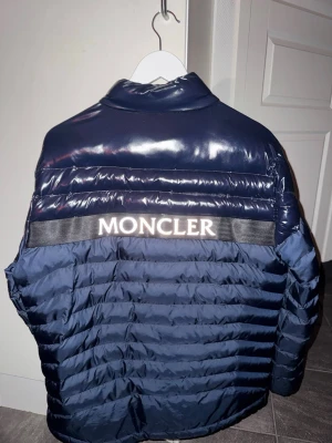 Mörkblå dunjacka från Moncler - Snygg mörkblå dunjacka från Moncler med glansig finish och quiltad design. Jackan har hög krage, dragkedja framtill, två sidofickor med dragkedja och stor Moncler-logga på ryggen. Perfekt för kalla vinterdagar och riktigt trendig look.