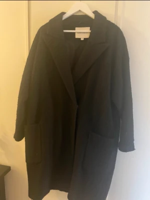Svart oversized kappa från Carin Wester - Stilren svart oversized kappa från Carin Wester  med klassisk krage och stora framfickor. Kappan har rak passform och är tillverkad i ett mjukt material som ger en avslappnad look. Perfekt för kyliga dagar och enkel att matcha med olika outfits.