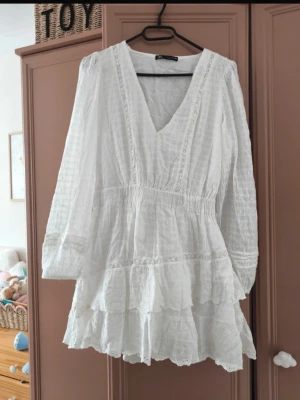 Zara  Ny söt Vit kort klänning med volang - Söt vit kortklänning med v-ringning och långa ärmar. Klänningen har volangdetaljer och spets längs sömmarna, vilket ger en romantisk känsla. Tillverkad i ett lätt, luftigt material som passar perfekt till varmare dagar.