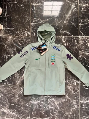 Nike Brasil vindjacka GORE-TEX - Ljusgrön vindjacka från Nike med Brasil-logga och flera sponsortryck som Guaraná Antarctica, Vivo och Itaú. Jackan har huva, hel dragkedja och är tillverkad i GORE-TEX-material som skyddar mot vind och regn. Perfekt för dig som gillar sportig stil.