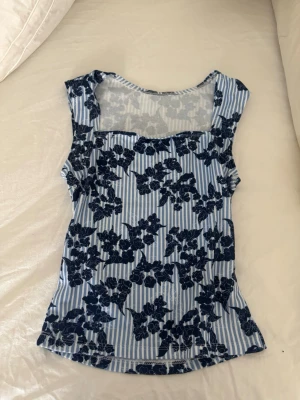Blommig blåvit ärmlös topp - Snygg ärmlös topp med blå och vita ränder och mörkblått blommönster. Toppen har en rak, något figurnära passform och fyrkantig ringning. Perfekt för dig som gillar en fräsch och somrig stil.