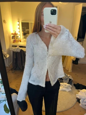 Vit topp med vida ärmar - Säljer en vit tröja/topp med v-ringning och vida ärmar. Tröjan har pärlknappar framtill och är gjord i ett lätt transparent material med diskret blommönster. Perfekt för dig som gillar trendiga detaljer och en luftig look.