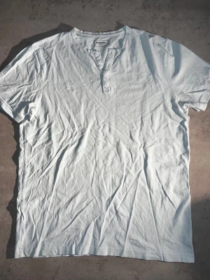 Ljusblå kortärmad t-shirt med knappar - L - Säljer en ljusblå kortärmad t-shirt från Dressmann med v-ringning och knappdetaljer framtill. Tillverkad i mjuk bomull som känns skön mot huden. Enkel och clean design som passar till det mesta. Perfekt för dig som gillar en klassisk och stilren look.