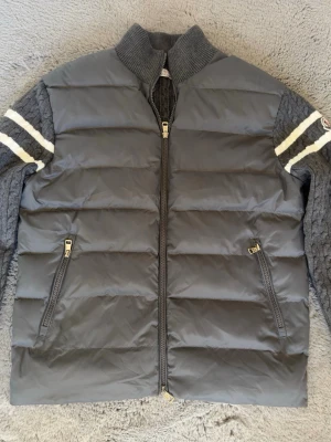 Grå Moncler pufferjacka med stickade ärmar - Snygg grå pufferjacka från Moncler med stickade ärmar och rygg i kabelstickat mönster. Jackan har vita ränder på ärmarna, ribbad krage och två dragkedjefickor fram. Perfekt för kalla vinterdagar och riktigt trendig look.