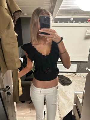 Svart croppad volangtopp - Säljer en svart croppad topp med volangdetaljer och djup v-ringning. Toppen är ärmlös och har en feminin, figurnära passform. Perfekt för dig som vill ha en trendig och snygg look.