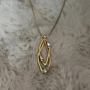 Ioaku Bliss halsband - Unikt halsband i guld från Ioaku med ett abstrakt, dubbelt lövformat hänge. Hänget är dekorerat med tre droppformade, klara stenar som ger extra bling. Kedjan är tunn och smidig, perfekt för att lyfta vilken outfit som helst.