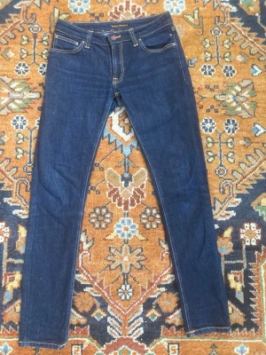 Mörkblå skinny jeans från Nudie - Snygga mörkblå skinny jeans från Nudie med klassiska kontrastsömmar och fem fickor. Jeansen har en smal passform och är tillverkade i stretchigt denimtyg för extra komfort. Bakfickorna har Nudies ikoniska broderi. Nästan helt nytt skick och knappast använda. S 29/32. Fråga om bilder och mått. Tar gärna prisrekomendationer!
