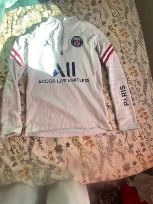 Vit PSG träningströja långärmad - Snygg vit långärmad träningströja från PSG med blå logga och text, röda och blå ränder på ärmarna samt halv dragkedja fram. Tillverkad i funktionsmaterial som passar perfekt till fotbollsträning eller sport. Paris-tryck på ena ärmen och Visit Rwanda på ryggen.