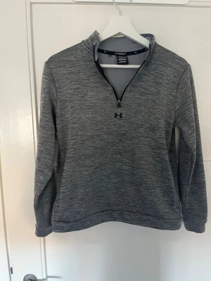 Golf tröja Grå halvzip tröja från Under Armour  - Snygg gråmelerad halvzip tröja från Under Armour med hög krage och lång ärm. Tillverkad i ett mjukt funktionsmaterial som passar perfekt för lager-på-lager. Diskret logga på bröstet och sportig look.