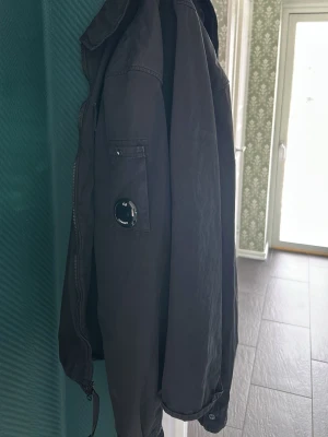 Svart vindjacka från C.P. Company - Säljer en svart vindjacka från C.P. Company med ikonisk linsdetalj på ärmen. Jackan har dragkedja framtill, hög krage och en ficka på ärmen. Materialet är tunt och smidigt, perfekt för blåsiga dagar.