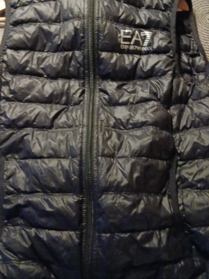 Svart dunväst från EA7 Emporio Armani - Snygg svart dunväst från EA7 Emporio Armani med quiltad design och dragkedja framtill. Västen har en diskret logga på bröstet och är lätt vadderad för extra värme. Perfekt att slänga över en hoodie eller tröja för en sportig look.