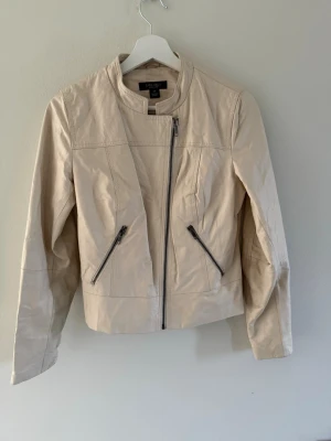 Beige skinnjacka från Sara Kelly - Snygg beige skinnjacka från Sara Kelly med cool asymmetrisk dragkedja och två snedställda fickor med dragkedja. Storlek 38 men skulle säga att den är liten i storlek.