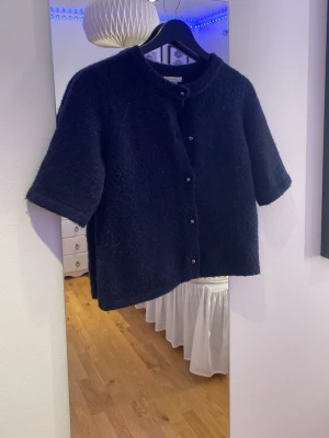 Mörkblå stickad kofta Gina Tricot - Säljer en mörkblå stickad kofta från Gina Tricot med korta ärmar och rund hals. Koftan har knappar framtill och är gjord i ett mjukt, fluffigt material. Perfekt att slänga över en topp för en chill look.