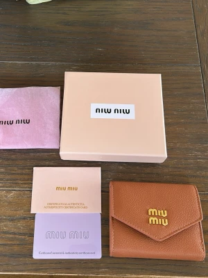 Brun plånbok från Miu Miu i läder - Säljer en snygg brun plånbok från Miu Miu i äkta läder med guldfärgad logga framtill. Plånboken har en stilren, vikbar design och kommer med originalask, dustbag och äkthetskort. Perfekt för dig som vill ha något lyxigt och trendigt i väskan.