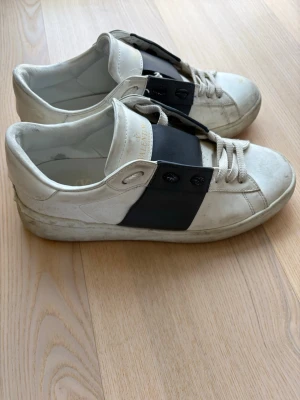 Valentino open - Snygga vita sneakers från Valentino med ett brett svart band över vristen och dekorativa nitar. Skorna har klassisk låg profil, vita snören och är tillverkade i läder. Perfekta för dig som vill ha en stilren men ändå unik look.