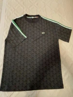 Mönstrad svart t-shirt från Lacoste - Snygg svart t-shirt från Lacoste med geometriskt mönster i grått. T-shirten har rund halsringning och gröna samt vita detaljer vid krage och ärmslut. Klassisk Lacoste-logga på bröstet. Perfekt för dig som gillar stilrena men unika plagg.