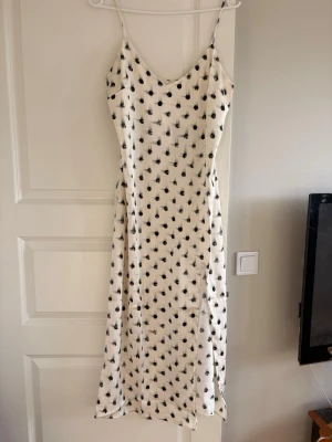 Vit långklänning med svart prickigt mönster - Snygg vit långklänning med svarta prickar och smala axelband. Klänningen har en slits framtill och är figurnära med en lätt v-ringad halsringning. Perfekt för sommarens alla tillfällen.