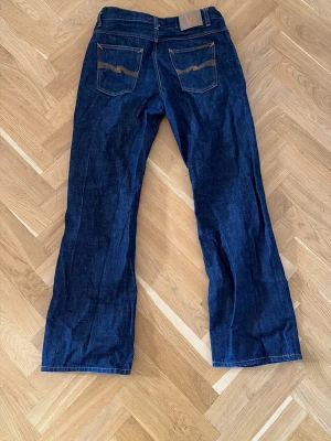 Mörkblå bootcut jeans - Säljer ett par mörkblå bootcut jeans från den populära modellen ”Nudie Jeans flare Glenn” i klassisk denim med kontrastsömmar och två bakfickor med vågig detalj. Jeansen har hög midja och är mer ”flare” nedtill. Storlek är 33 i waist & 34 i längd. Använda i cirka 6 månader och är i ett bra skick! Skicka meddelande om ni har några funderingar.