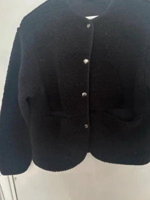 Svart teddyjacka med tryckknappar - Mysig svart teddyjacka med rund halsringning och silverfärgade tryckknappar framtill. Jackan har två fickor och är gjord i fluffigt teddy-material som ger en trendig look. Perfekt för att hålla värmen under kyliga dagar.