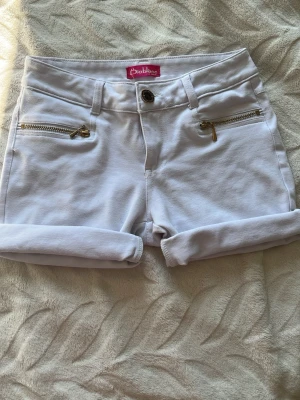 Vita shorts - Snygga vita shorts med uppvikta ben och två guldfärgade dragkedjor framtill. Klassisk femficksmodell med bälteshällor och knappstängning. Perfekta för sommaren och har en fräsch, clean look. Köpte dessa secondhand och klippte dom själv.