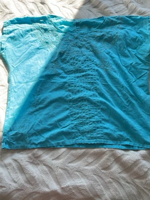 Blå blus med broderi - Klarblå blus i luftig bomull med fina broderade detaljer framtill. Blusen har rak passform och korta ärmar, samt knäppning bak i nacken. Perfekt för dig som gillar färg och enkel stil.
