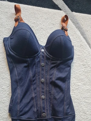 Mörkblå denim body med knappar - Snygg mörkblå body i denim-look med markerade sömmar och dekorativa knappar framtill. Kupor med bygel och justerbara axelband med läderdetaljer. Materialet ser ut som stretchig jeans, vilket ger en cool och trendig känsla.