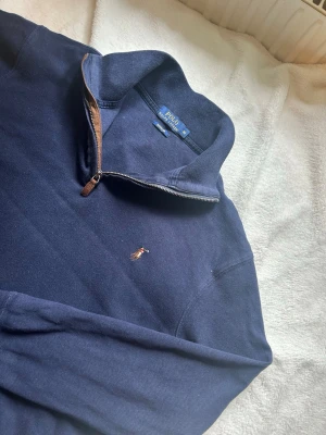 Mörkblå halvzip från Polo Ralph Lauren - Mörkblå långärmad tröja från Polo Ralph Lauren med halv dragkedja och klassisk broderad logga på bröstet. Tröjan har ståkrage med bruna detaljer vid dragkedjan och är tillverkad i mjukt bomullsmaterial.