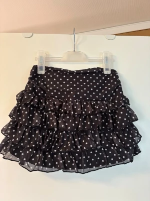 Svart volangkjol med vita prickar - Söt svart kortkjol med vita prickar och flera lager volanger. Kjolen har en luftig och lätt känsla, perfekt för dig som gillar en lekfull stil. Inbyggda shorts. De är helt nya och oanvända!