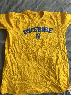 Gul Sverige t-shirt med tryck - Gul t-shirt med blå texten 'Sverige' och svensk flagga på bröstet. Klassisk rund hals och korta ärmar. Perfekt för att visa ditt stöd för Sverige. Tillverkad i mjuk bomull för skön känsla.