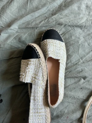 Beige loafers med pärldetaljer - Säljer ett par snygga beige loafers med svart tåparti och flätad sula. Ovandelen är i ett tweed-liknande tyg med små pärlor insydda för extra stil. Skorna har en klassisk slip-on design och passar perfekt till jeans eller kjol.