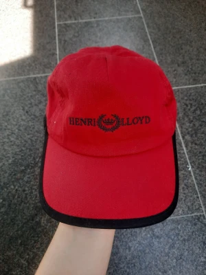 Röd keps Henri Lloyd - Fin röd keps av märket Henri Lloyd. Den går att justera i storlek med resårband. 