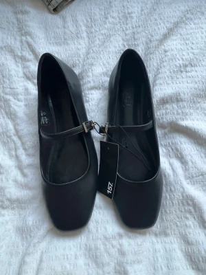 Svarta Mary Jane ballerinaskor - Stilrena svarta ballerinaskor med klassisk Mary Jane-rem och silverfärgat spänne. Skorna har fyrkantig tå och låg klack, vilket ger en modern touch. Tillverkade i ett slätt material som ger en clean look.