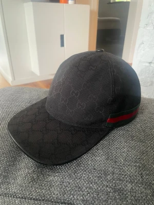 Svart Gucci keps - Snygg svart keps från Gucci med det klassiska GG-mönstret över hela kepsen. På sidan finns en grön och röd rand som ger en exklusiv touch. Justerbar rem baktill för perfekt passform. Perfekt accessoar för dig som gillar streetwear och lyx.