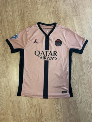 PSG rosa matchtröja med svarta detaljer - Säljer en snygg rosa PSG matchtröja med svarta detaljer och vertikal rand. Tröjan har korta ärmar, V-ringning och Paris Saint-Germain-logga på bröstet. Tillverkad i lätt och ventilerande material, perfekt för fotboll eller träning. Passar dig mellan 170-180