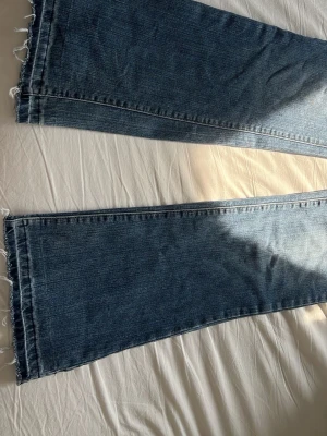 Blåa bootcut jeans med rå kant - Säljer ett par blåa bootcut jeans med trendig rå kant nertill. Jeansen har en klassisk tvätt och breda ben, vilket ger en avslappnad och cool vibe. Perfekta för dig som gillar en lite mer edgy stil. Materialet är klassisk denim.