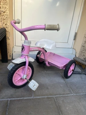 Rosa trehjuling med vita detaljer - Säljer en söt rosa trehjuling med vita handtag, pedaler och skärm. Sätet är vitt och ramen är i metall. Trehjulingen har svarta däck och en liten plattform baktill. Perfekt för yngre barn som vill lära sig cykla.