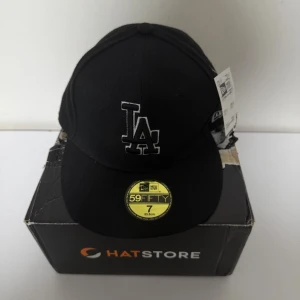 New Era LA 59Fifty Cap - Size 7 (55,8cm) - Kepsen är helt ny, inga skador eller slitage och tagsen sitter kvar.  Klassisk New Era 59FIFTY fitted-keps med den ikoniska LA-loggan i svart med vit kant. Stilren design som passar perfekt till alla outfits.  Storlek: 7 (55,8 cm) Rabatt vid köp av flera plagg🏷️
