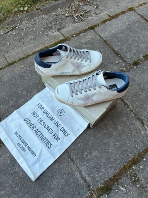 Golden Goose vita sneakers med blå detalj - Golden Goose sneakers i vitt läder med den ikoniska stjärnan på sidan och mörkblå hälkappa. Skorna har grå skosnören och en klassisk låg siluett. Snygg och avslappnad design som passar perfekt till jeans eller shorts.