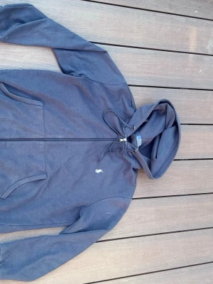 Mörkblå hoodie från Polo Ralph Lauren - Mörkblå hoodie med dragkedja från Polo Ralph Lauren. Klassisk design med huva och dragsko, två fickor framtill och den ikoniska broderade loggan på bröstet. Tillverkad i mjukt bomullsmaterial som är skönt att bära.