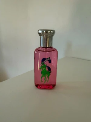 Ralph Lauren Big Pony Parfum - En snygg parfymflaska i rosa glas från Ralph Lauren med silverfärgat lock. Flaskan har en stor grön logga med en polospelare och en blå siffra på framsidan. Perfekt som en färgglad detalj på sminkbordet.