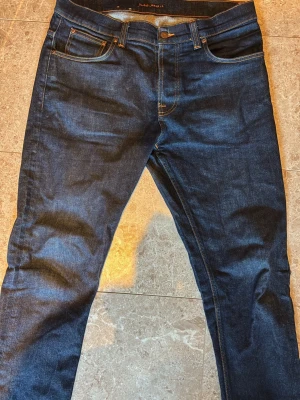 Nudie Jeans - Grim Tim - W36 L32 - Snygga mörkblå jeans med raka ben och klassisk femficksmodell. Jeansen har kontrastsömmar och är tillverkade i ett slitstarkt denimtyg. Passar perfekt till en avslappnad stil och funkar året runt.
