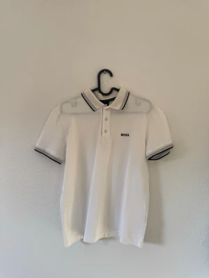 Vit piké från BOSS  - Klassisk vit pikétröja från BOSS med svarta detaljer på krage och ärmslut. Tröjan har korta ärmar, knappslå framtill och en stilren logga på bröstet. Perfekt för en clean och enkel look.