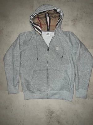 Grå zip hoodie med mönstrad huva - Snygg grå zip hoodie med dragkedja och fickor framtill. Huvan har ett klassiskt rutigt mönster på insidan och justerbara snören. På bröstet finns en broderad logga i vitt. Perfekt för en chill och stilren look.