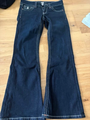 Mörkblå bootcut jeans med kontrastsömmar - Snygga mörkblå bootcut jeans med vita kontrastsömmar och klassisk femficksdesign. Jeansen har låg midja och vida ben, perfekta för en retroinspirerad look. Materialet är kraftig denim som ger en schysst passform.