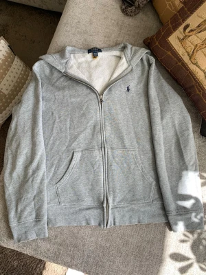 Grå hoodie från Polo Ralph Lauren - Säljer en grå zip-hoodie från Polo Ralph Lauren med blå logga på bröstet. Hoodien har dragkedja, två fickor fram och mjukt vitt foder på insidan av huvan. Perfekt för chill dagar och enkel att matcha med jeans eller joggers. Storlek 18-20 år, motsvarar vuxen S.