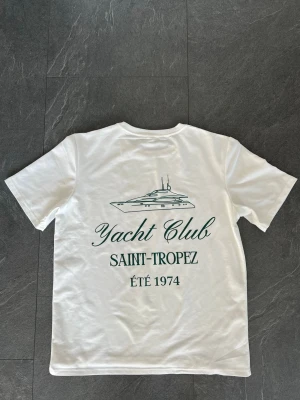 Vit Yacht Club Saint-Tropez t-shirt - Säljer en vit t-shirt med tryck av en yacht och texten 'Yacht Club Saint-Tropez ÉTÉ 1974' i grönt på både fram- och baksidan. T-shirten har rund hals och korta ärmar. Perfekt för dig som gillar clean stil med marina vibbar.