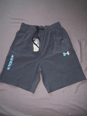Gråa under armour shorts med reflexdetaljer - Snygga svarta shorts med elastisk midja och snörning. Reflexdetaljer i regnbågsfärger på ena benet och logga på andra sidan. Perfekta för träning eller chill. Lätta och bekväma med sportig vibe.