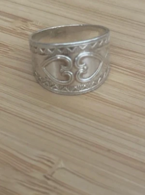 Bred silverfärgad ring med mönster  - Säljer en västerbottensring bred ring i silverfärg med detaljerat reliefmönster och hjärtformade inslag. Behövs putsas lite. Storlek 2.5 cm ish diameter. Ringen har dekorativa kanter med små trianglar och en blank finish. Perfekt för dig som gillar unika accessoarer med boho-vibe. 